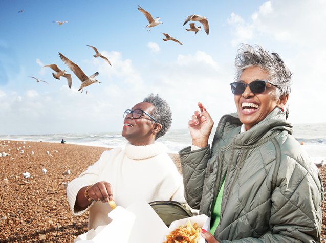 Two Black women laughing and eating fish and chips on a pebble beach while seagulls swoop around them. 